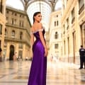 A woman wearing the elegant NC1049, a floor-length, off-the-shoulder purple gown, stands in a sophisticated glass-domed building reflecting classical architecture. Her dark hair is styled neatly as she glances over her shoulder with an air of confidence and grace. Other people can be seen in the background. Angels Formal Wear