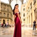 A woman in a flowing, floor-length NC1049 red gown stands confidently in an ornate, sunlit gallery with a glass-domed ceiling and columns. She faces the camera, showcasing the off-the-shoulder sleeves of her dress. Her long brown hair cascades down her back, complemented by large hoop earrings. In the background, people are walking through the space. Angels Formal Wear