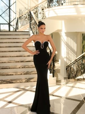 A woman in the LUNA NC1052 evening gown, featuring a strapless design and heart-shaped neckline, stands at the base of a grand marble staircase adorned with intricate wrought iron railings. With her hair styled in a high bun, she confidently poses in this well-lit luxurious interior, showcasing selections from an exclusive online dress shop on the Sunshine Coast. Angels Formal Wear