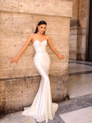 A person wearing the NC1054, a strapless, form-fitting, white evening gown, poses elegantly against a stone wall. They have long, dark hair, bold makeup, and large statement earrings. The floor-length dress flares slightly at the bottom. The background is an old building—perfect for any special occasion and available at Dress Shop Sunshine Coast with Afterpay options. Angels Formal Wear