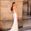 A woman stands elegantly with her back slightly turned, wearing the NC1054, a strapless, floor-length, shiny white gown from Dress Shop Sunshine Coast. Her hand rests on a textured stone wall, and she has long, sleek hair. She accessorizes with large, circular earrings and exudes a confident, poised demeanor. Angels Formal Wear