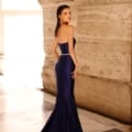 A woman stands in an elegant stone building, wearing the form-fitting, strapless deep blue NC1054 gown from Dress Shop Sunshine Coast, which features a long train. She glances over her shoulder with her dark hair pulled back and accessorized with large circular earrings. The lighting beautifully highlights the gown’s sheen. Afterpay available! Angels Formal Wear