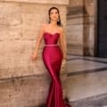 A woman stands leaning against a stone wall in an elegant, form-fitting, strapless magenta gown (Product Name: NC1054) that flows to the floor with a mermaid silhouette. Large, intricate earrings complement her evening wear. The background features a marble floor and architectural columns reminiscent of the Sunshine Coast's opulence. Angels Formal Wear