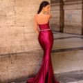 A woman stands with her back to the camera, wearing an NC1054 gown - a long, glossy, burgundy strapless dress featuring a small train. Positioned against a stone wall in an elegant architectural setting reminiscent of the Sunshine Coast, her long dark hair is styled straight. Showcasing stunning evening wear, she touches her face with one hand. Angels Formal Wear