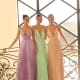 Three women stand side by side in elegant ROMA NC1060 floor-length dresses with deep V-necklines. The pastel-colored gowns—one in lavender, one in mint green, and one in light peach—evoke the serene beauty of the Sunshine Coast. Indoors, next to a decorative railing and a large window, they smile and pose together. Angels Formal Wear