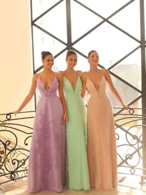 Three women stand side by side in elegant ROMA NC1060 floor-length dresses with deep V-necklines. The pastel-colored gowns—one in lavender, one in mint green, and one in light peach—evoke the serene beauty of the Sunshine Coast. Indoors, next to a decorative railing and a large window, they smile and pose together. Angels Formal Wear