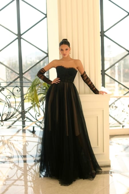 A woman stands in an elegant room with large window panes behind her, reminiscent of the panoramic views of Sunshine Coast. She is wearing NC1064, a strapless, floor-length black gown accented with sheer details and paired with long matching black gloves. Her hair is styled in a sleek updo. Angels Formal Wear