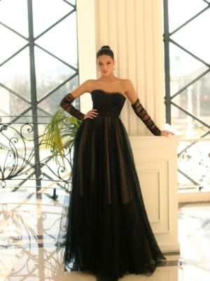 A woman stands in an elegant room with large window panes behind her, reminiscent of the panoramic views of Sunshine Coast. She is wearing NC1064, a strapless, floor-length black gown accented with sheer details and paired with long matching black gloves. Her hair is styled in a sleek updo. Angels Formal Wear