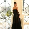 A woman stands in an elegant setting with large windows and decorative metal railings. She is wearing the NC1064, a black, off-the-shoulder, sheer gown from an online dress shop, and has her hair styled in a bun. She looks over her shoulder towards the camera with a serene expression, capturing the essence of Sunshine Coast charm. Angels Formal Wear