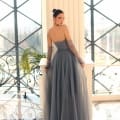 A woman stands inside a modern building with large windows, wearing the NC1064 sleeveless, flowing gray evening gown from an online dress shop. Her hair is styled in a sleek updo as she rests one hand on a white pillar. Sunlight streams through the windows, illuminating the scene like a day on the Sunshine Coast. Angels Formal Wear