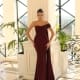 A woman stands elegantly in an opulent room at Dress Shop Sunshine Coast, wearing a floor-length, off-the-shoulder maroon gown (Product Name: NC1065). Her hair is styled in a sleek updo, and she accessorizes with large hoop earrings. The backdrop features tall windows with black framing, letting in ample natural light. Angels Formal Wear