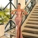 A woman confidently stands in front of an elegant staircase, graced by the TAMARA NC1074, a silky pink evening gown with a high slit. Her hands rest on her hips as she complements the look with white high heels. Sunlight streams through large windows, casting a Sunshine Coast glow on the scene. Angels Formal Wear