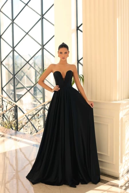 A woman wearing a long, elegant, strapless black gown (Product Name: NC1075) stands in an opulent setting with tall white columns and large lattice windows. She has her hair tied back in a sleek bun and is holding the sides of her dress from Dress Shop Sunshine Coast, displaying its flowing fabric. Angels Formal Wear