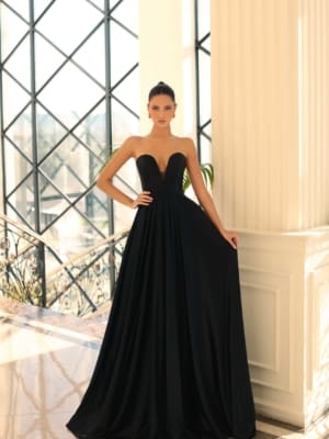 A woman wearing a long, elegant, strapless black gown (Product Name: NC1075) stands in an opulent setting with tall white columns and large lattice windows. She has her hair tied back in a sleek bun and is holding the sides of her dress from Dress Shop Sunshine Coast, displaying its flowing fabric. Angels Formal Wear