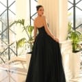 A woman stands gracefully in a sunlit room with large windows and white columns, evocative of the elegant ambiance at Dress Shop Sunshine Coast. She is adorned in the long, strapless black gown NC1075 and looks over her shoulder. Green potted plants flank her on either side, adding a touch of nature to the setting. Angels Formal Wear