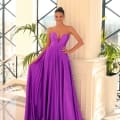 A person stands confidently in a brightly lit, elegant room with large windows and greenery. They are wearing a strapless, flowing purple gown (Product Name: NC1075) from Dress Shop Sunshine Coast and large hoop earrings, with their hair styled up. They pose with one hand resting on a white column. Angels Formal Wear