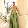 A woman stands gracefully in a well-lit room with large windows, wearing an elegant, strapless green gown (Product Name: NC1075) from Dress Shop Sunshine Coast. The V-shaped neckline leads to a dress that flows to the floor, fanning out around her feet as she rests one hand on a white pillar, gazing confidently ahead. Angels Formal Wear