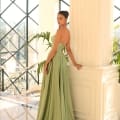 A young woman in a flowing, backless NC1075 green dress stands beside a white column in a spacious, sunlit room with large windows and a view of ornate railings and plants. She looks over her shoulder towards the camera, with her hair pulled back in an elegant updo—a vision from any Dress Shop on the Sunshine Coast. Angels Formal Wear