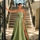 A woman stands at the bottom of an elegant staircase, wearing a strapless, flowing green ANNA NC1076 gown from a local Sunshine Coast dress shop. She poses with one hand on the banister and the other on the door frame in a well-lit, luxurious setting with intricate railing designs. Angels Formal Wear