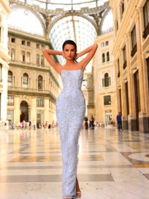 A woman in a chic, figure-hugging NC1082 gown in light blue with intricate silver embroidery poses gracefully with her hands behind her head. She stands in a magnificent indoor setting featuring a glass dome ceiling and elegant arched walkways. Complementing her look, she wears high heels and statement earrings. Angels Formal Wear