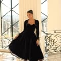 A woman wearing a black, strapless velvet dress with a matching bolero smiles as she poses in front of a large window with an iron railing. In the elegant room NC1084, marble floors and classical architecture, including tall columns, enhance the timeless beauty of the scene. Angels Formal Wear