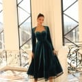 A woman stands in a spacious, elegant interior with large windows and ornate black railings. She wears the NC1084, a dark green velvet dress featuring a sweetheart neckline and long sleeves, paired with strappy heels. Her hair is styled in a sleek, high ponytail. Angels Formal Wear