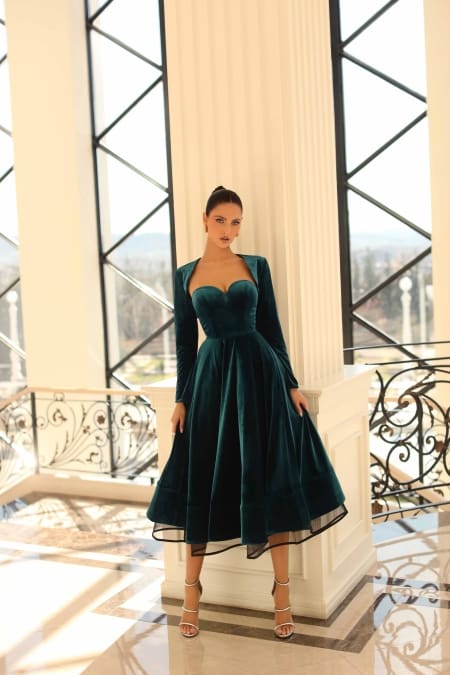 A woman stands in a spacious, elegant interior with large windows and ornate black railings. She wears the NC1084, a dark green velvet dress featuring a sweetheart neckline and long sleeves, paired with strappy heels. Her hair is styled in a sleek, high ponytail. Angels Formal Wear