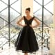 A woman in a black, off-the-shoulder, tea-length NC1089 dress from the local Dress Shop stands indoors by a decorative black wrought iron railing. She is smiling and posing with her hands on her hips. Sunlight streams in through large windows with a diamond-shaped grid pattern behind her. Angels Formal Wear