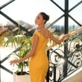 A woman wearing a yellow dress stands on a staircase by a large window. She is facing slightly away from the camera, her hand resting on the NC1091 railing with a potted plant nearby. Sunlight streams in, casting a glow over the scene. Angels Formal Wear