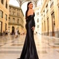 A woman in a sophisticated, black floor-length NC1093 dress exudes confidence as she poses in an elegant, arched walkway adorned with intricate architectural details. Smiling radiantly, with long earrings and a sleek, slicked-back hairstyle, she perfectly embodies the timeless allure of the NC1093 dress in this breathtaking moment. Angels Formal Wear