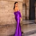 A woman stands against a stone wall, adorned in a vibrant NC1093 purple off-the-shoulder gown that enhances her figure and cascades gracefully to the floor. Her long, straight hair complements her look, accessorized with large gold earrings. The setting seems urban and sophisticated. Angels Formal Wear