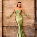 A woman stands against a stone wall wearing the NC1093, a form-fitting, strapless, shimmery green gown with a mermaid silhouette. She accessorizes with matching green gloves that extend above her elbows and large hoop earrings. Her long hair flows down her back. Angels Formal Wear