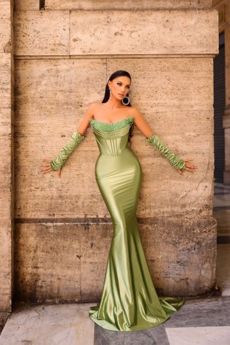 A woman stands against a stone wall wearing the NC1093, a form-fitting, strapless, shimmery green gown with a mermaid silhouette. She accessorizes with matching green gloves that extend above her elbows and large hoop earrings. Her long hair flows down her back. Angels Formal Wear