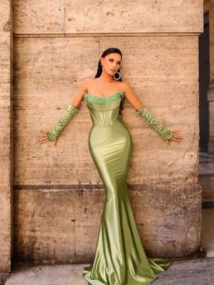 A woman stands against a stone wall wearing the NC1093, a form-fitting, strapless, shimmery green gown with a mermaid silhouette. She accessorizes with matching green gloves that extend above her elbows and large hoop earrings. Her long hair flows down her back. Angels Formal Wear