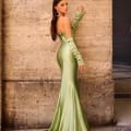 A woman stands against a rough-textured stone wall, wearing an elegant, form-fitting satin green dress with off-the-shoulder long sleeves. Her long dark hair is accessorized with large hoop earrings as she gazes over her shoulder at the camera. The scene is identified by the intricate marble floor patterns in the backdrop, known as NC1093. Angels Formal Wear