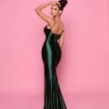 A woman stands against a pink background, wearing the NP144 Deep floor-length forest green satin dress. She has her hair in a high bun and looks back over her shoulder with a slight smile, one hand resting on her head. This elegant look could be right out of the Best Dress Shop on the Sunshine Coast. Angels Formal Wear