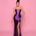 A woman stands against a pink background, wearing the NP144 Deep, a form-fitting, floor-length dark purple satin dress with a high slit from the Best Dress Shop on the Sunshine Coast. She poses with one hand on her hip and the other resting down. She has an updo hairstyle and wears hoop earrings and high heels. Angels Formal Wear