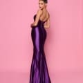 A woman in a shiny, fitted purple evening gown called NP144 Deep from the best dress shop on the Sunshine Coast stands against a pink background. Her stunning gown features a strapless, mermaid silhouette. She has an elegant, high bun hairstyle and is posing with her back slightly turned and hands on her waist, looking over her shoulder. Angels Formal Wear