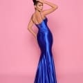 A woman stands against a pink background, dressed in a backless, floor-length, royal blue satin gown named NP144 Deep from the Best Dress Shop. Her hair is styled in an elegant updo, and she poses with one hand touching her head and the other resting on her hip, showcasing the dress’s design from the back. Angels Formal Wear