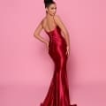 A woman stands against a pink background, wearing the NP144 Deep, a backless, form-fitting, floor-length red satin gown from the Best Dress Shop. Her hair is styled in an elegant updo. She poses with one hand on her hip, looking over her shoulder. Angels Formal Wear