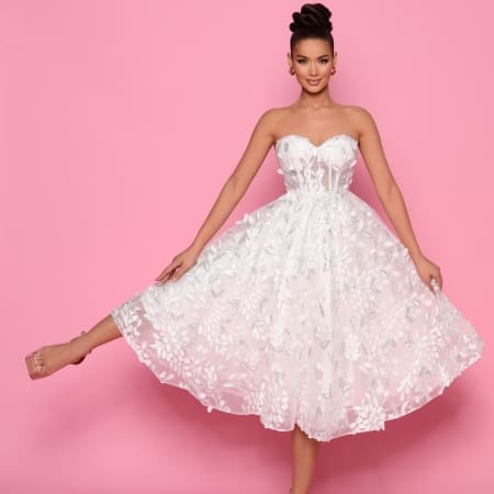 A woman in the NP145 strapless, white, floral lace dress from the Best Dress Shop poses against a pink background. She is smiling and lifting one leg slightly behind her, showcasing her nude high-heeled shoes. Her hair is styled in an elegant updo, reminiscent of the timeless style often seen on the Sunshine Coast. Angels Formal Wear