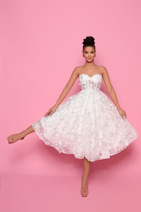 A woman in the NP145 strapless, white, floral lace dress from the Best Dress Shop poses against a pink background. She is smiling and lifting one leg slightly behind her, showcasing her nude high-heeled shoes. Her hair is styled in an elegant updo, reminiscent of the timeless style often seen on the Sunshine Coast. Angels Formal Wear