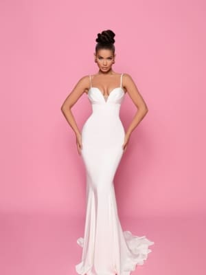 A woman poses against a pink background wearing a fitted white gown with a plunging neckline and thin straps, purchased from the Cocktail Dress Shop and identified as product NP146. Her hair is styled in a high updo, and she stands with hands resting on her hips, emphasizing the dress's elegant, flowing design. Angels Formal Wear