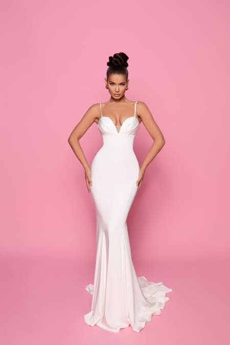 A woman poses against a pink background wearing a fitted white gown with a plunging neckline and thin straps, purchased from the Cocktail Dress Shop and identified as product NP146. Her hair is styled in a high updo, and she stands with hands resting on her hips, emphasizing the dress's elegant, flowing design. Angels Formal Wear