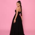 A woman in NP147 stands against a pink background. Her strapless black gown, ideal for Race Day Dresses, boasts a corset-style lace-up back. She has long, dark hair styled in an elegant braid and wears noticeable hoop earrings. She gazes over her shoulder towards the camera. Angels Formal Wear