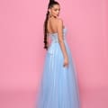 A woman with long, braided hair is posing against a pink background on the Sunshine Coast. She is wearing product NP147, a strapless, floor-length, light blue gown featuring a lace-up back and intricate bodice details. With one hand resting on her hip, she looks over her shoulder, exuding elegance perfect for race day dresses. Angels Formal Wear