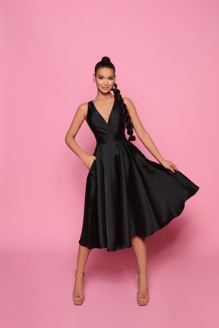 A woman poses against a pink background, wearing the ELORA NP150, a black sleeveless knee-length cocktail dress with a deep V-neck and pleated skirt from Sunshine Coast's finest Cocktail Dress Shop. She has long, braided hair and is holding the sides of her dress while smiling. She is accessorized with large hoop earrings and nude high-heeled shoes. Angels Formal Wear