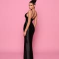 A woman with hair styled in a high bun is standing against a pink background, looking over her shoulder at the camera. She is wearing the floor-length, black, sleeveless EVIE NP159 gown with thin straps and a side slit—perfect for cocktail dress enthusiasts on the Sunshine Coast. Angels Formal Wear