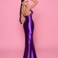 A woman with long braided hair poses against a pink background, wearing the EVIE NP159, a long, form-fitting, shiny purple cocktail dress with thin straps. She stands with one hand on her head, looking over her shoulder towards the camera. This chic look is perfect for summer nights in Sunshine Coast. Angels Formal Wear