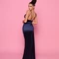 A woman stands on a pink background, wearing the elegant NP162 dress in navy blue, featuring a backless design and thin straps. Her hair is styled in an elegant updo as she looks over her shoulder towards the camera with a confident expression. This stunning ensemble is from the Best Sunshine Coast Dress Shop. Angels Formal Wear